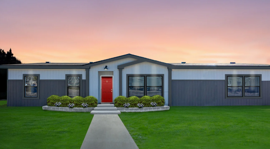 A modern manufactured home with a red door and well-kept landscaping at sunset