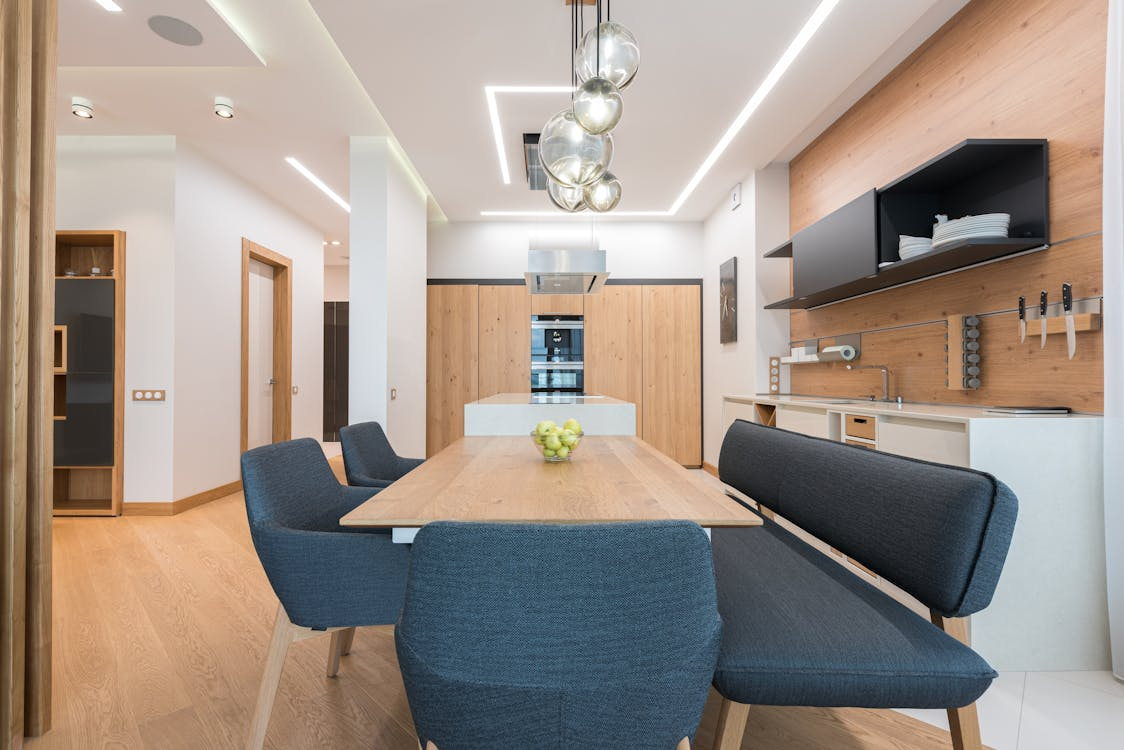 A sleek kitchen with wood cabinets, a built-in oven, dining area, and pendant lighting.